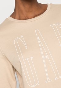 Felpa beige con scollo rotondo, caratterizzata da grandi scritte bianche. Il tessuto ha una texture liscia con dettagli di cuciture visibili.