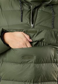 Green padded jacket with a zippered neck, two front pockets, and a hand partially tucked into the upper pocket. Features a smooth texture.