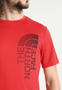 Camiseta roja de manga corta hecha de tejido transpirable, con un diseño de logo de puntos negros en la parte delantera. Cuello redondo con un ajuste relajado.