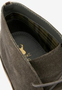 Zapato de ante marrón con una superficie texturizada, que presenta una plantilla negra y un patrón de cuadros en el interior, decorado con acentos cosidos.