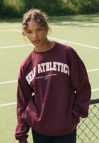 Burgundi värvi sviiter, millel on suur lõige ja valge, tekstuuriga "SELF ATHLETICS" logo ning tekst. Pehme kangas, ümmargune kaelus, madalad õlapiirkonnad.