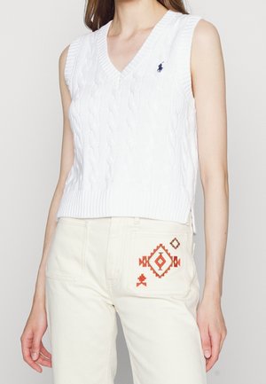 Kobieta ubrana w biały, bezrękawnik w splot warkoczowy oraz kremowe spodnie z czerwonym geometrycznym haftem na kieszeni, stojąca.