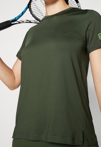 Oliwkowa sportowa koszulka z krótkim rękawem, okrągłym dekoltem, bocznymi rozcięciami, wykonana z gładkiego materiału, z logo na lewym rękawie.