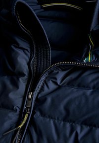Námořnická puffer bunda uvnitř s černým zipem a žlutou akcentovací šňůrou; prošívaná textura a hladká tkanina viditelné na otvoru.