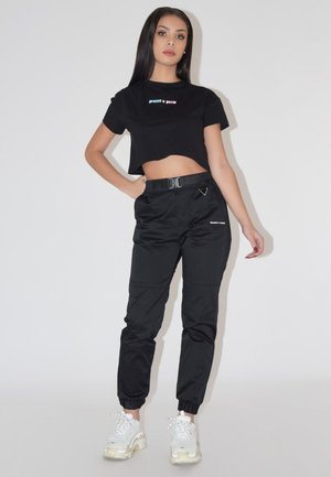 Donna con lunghi capelli scuri che indossa una maglietta nera corta e pantaloni da jogging neri, in piedi con una mano sul fianco e con scarpe da ginnastica bianche.