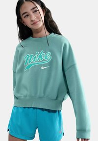 Giovane donna che indossa una felpa Nike turchese e pantaloni corti Nike blu, in piedi con una postura rilassata e leggermente inclinata contro uno sfondo semplice.