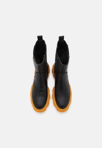 Une paire de bottines en cuir noir avec des semelles en caoutchouc épaisses et orange vif, des panneaux élastiques sur les côtés et des languettes de traction sur fond blanc.