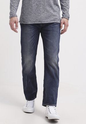Mann, der verblasste dunkelblaue Jeans, ein graues Langarmshirt und weiße Schnürsneaker trägt, steht vor einem schlichten hellen Hintergrund.