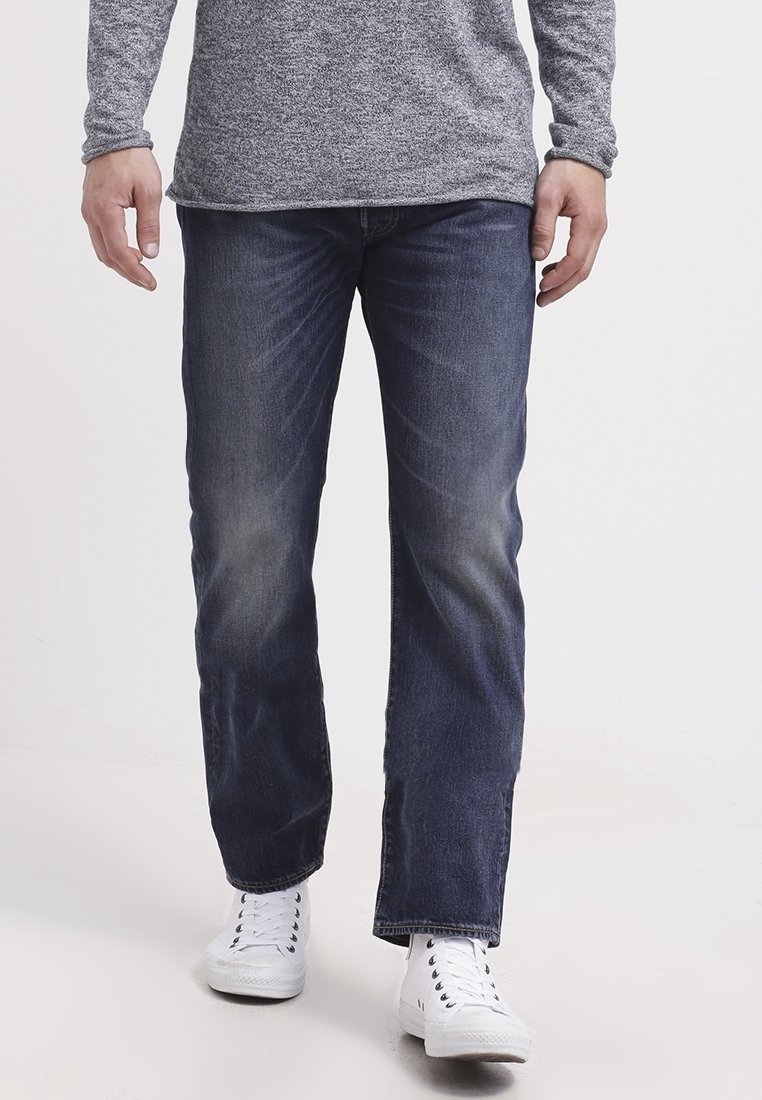 Homme portant un jean bleu foncé délavé, un t-shirt gris à manches longues et des baskets blanches à lacets, debout sur un fond clair uni.
