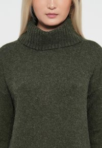 Mörkgrön stickad polotröja med ribbad krage och textilstruktur. Passformen är något avslappnad och har ett mjukt, fuzzy utseende.