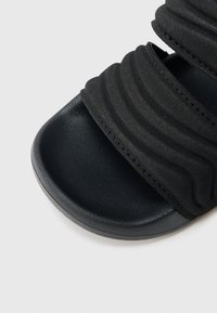 Mustad liugusandaalid, millel on pehmed, tekstureeritud sangad kogutud disainiga ja siledad, ümarad tallad. Materjal paistab kummine ja polsterdatud.