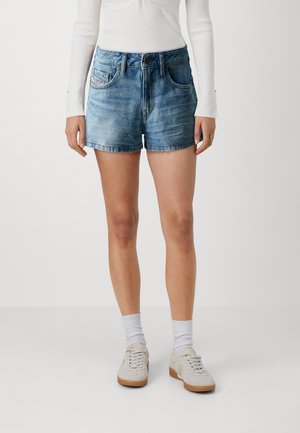 Femme portant un short en jean bleu, un haut blanc à manches longues, des chaussettes blanches et des baskets blanches, debout sur un fond uni.