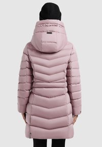 Kvinne iført en lyserosa quiltet dunjakke med hette og en svart strikket lue, vist fra ryggen mot en ensfarvet bakgrunn.