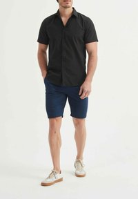 Chemise noire à manches courtes avec boutons, associée à un short bleu marine et des baskets blanches. La chemise a un col pointu ; le short est jusqu'aux genoux.