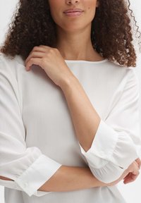 Blouse blanche en tissu texturé avec des manches larges et élastiquées. Présente des poignets boutonnés, un col rond et une coupe décontractée.