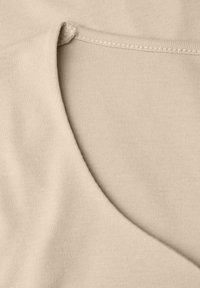 Zbliżenie beżowej tkaniny, miękka tekstura, z zaokrąglonym dekoltem i detalami szycia wzdłuż krawędzi, co tworzy gładkie przejście.