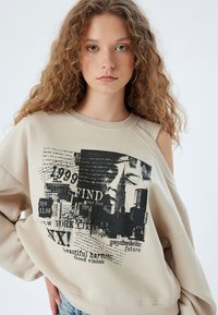 Felpa oversize beige con stampa grafica nera che presenta testo e design di skyline. Dettaglio con spalle scoperte, tessuto morbido.