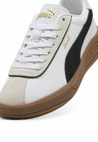 Chaussure de sport blanche avec une bande en suède noir, un logo doré et un embout beige, avec une tige texturée et une semelle en caoutchouc.