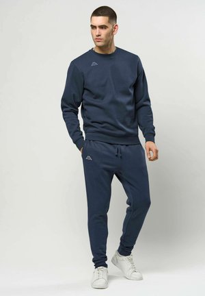 Młody mężczyzna stojący, ubrany w granatowy dres z logo, białe tenisówki, patrzący na bok na tle jednolitego szarego tła.