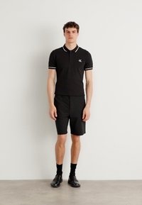 Svart kortermet polo-skjorte med hvite detaljer og logo, sammen med svarte shorts og sokker, iført svarte sko. Slitesterk og glatt tekstil.