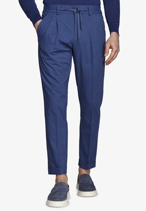 Hombre vistiendo pantalones azul ajustados con cintura de cordón, puños remangados, suéter azul marino y mocasines de ante azul con suelas blancas.