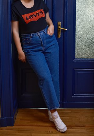 Personne appuyée contre une porte bleue, portant un t-shirt noir Levi's, un jean taille haute bleu et des baskets blanches sur un sol en bois.