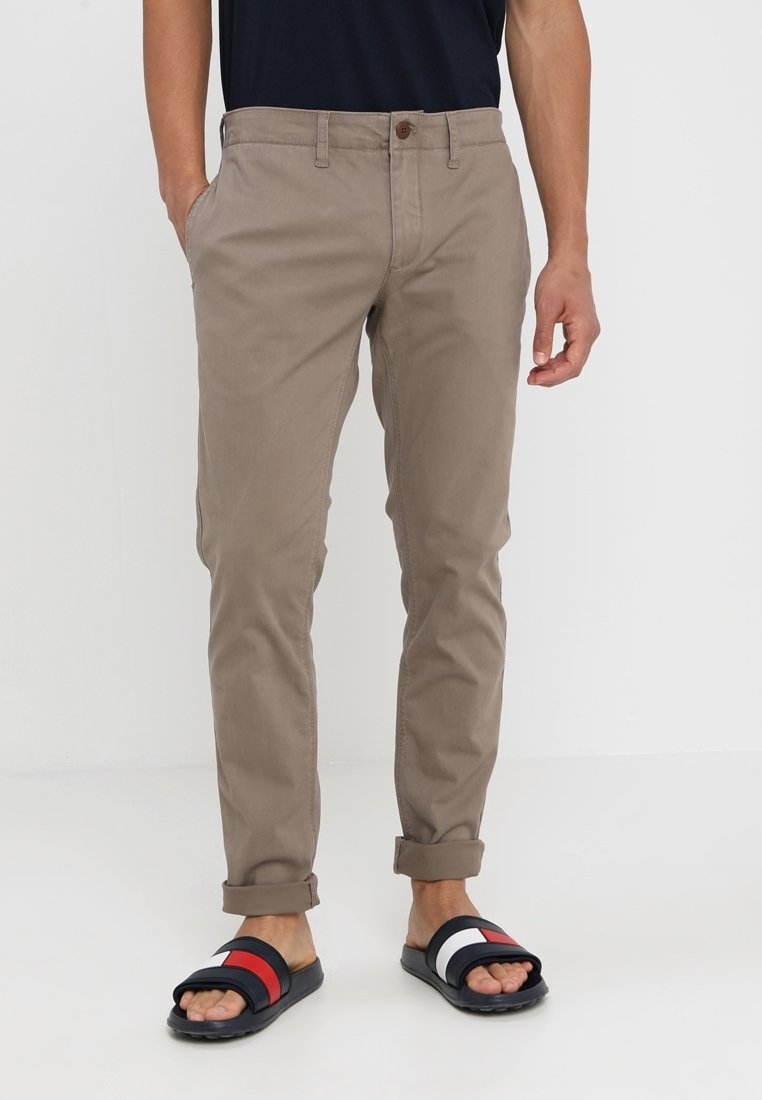 Jasnobrązowe chinosy o prostych nogawkach, z zapięciem na guzik i bocznymi kieszeniami. Noszone z czarno-białymi sandałami z kolorowymi akcentami.