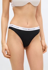 Γυναικείο σώμα που φοράει μαύρο μπικίνι σλιπ Tommy Hilfiger με λευκή ζώνη μέσης και μπεζ σουτιέν σε λευκό φόντο.