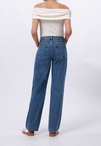 Žena stojící s rukama v zadních kapsách, oblečená v bílém topu odhalených ramen, vysokým pasem modrých džín a sandálech s leopardím vzorem na podrážkách.