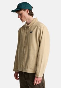 Μπεζ σακάκι από κορντόρι με γιακά, εμπρόσθιες ραφές και μικρό έμβλημα. Μακριά μανίκια με μανσέτες με κουμπιά, φορεμένο πάνω από σκούρο παντελόνι.