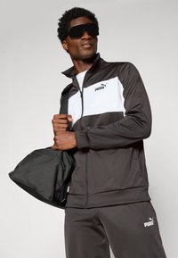 Man wearing black and white Puma tracksuit and large black sunglasses, holding a black Puma bag strap over his shoulder.