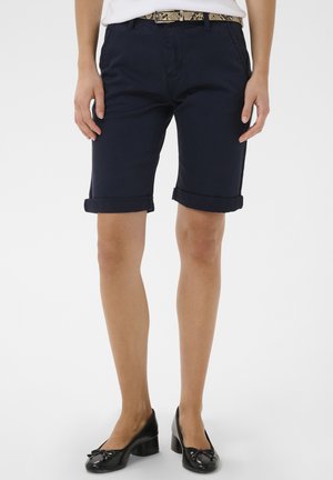 Kobieta w podwiniętych granatowych krótkich spodenkach, białym topie, pasku w wężowy wzór oraz czarnych butach na niskim obcasie, stojąca na jednolitym tle.