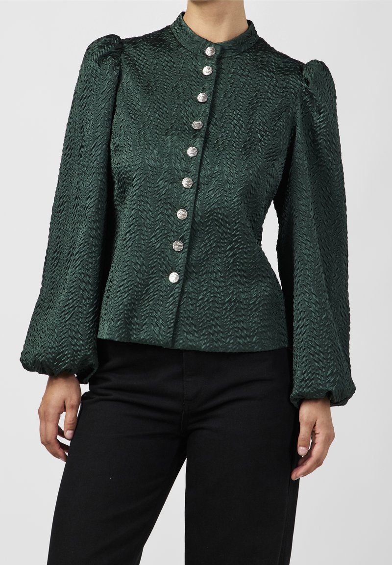 Femme portant un chemisier texturé vert foncé à manches longues avec des boutons argentés et un pantalon noir, debout devant un fond uni.