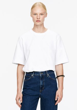 Weißes kurzärmliges T-Shirt mit lockerer Passform, kombiniert mit hochgeschnittenen dunkelblauen Jeans, dazu eine schlichte silberne Halskette und ein schwarzes Armband.