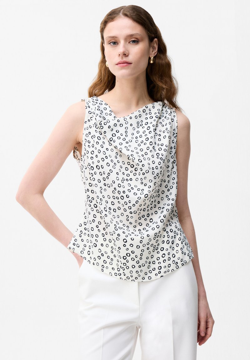 Jeune femme portant un chemisier blanc sans manches avec un motif de cercles noirs et un pantalon blanc, debout avec une main dans la poche.