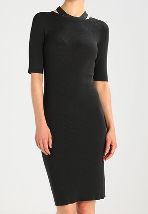 Vrouw draagt een nauwsluitende zwarte jurk tot op de knie met korte mouwen en een uitgesneden halsdetail, staand tegen een effen lichte achtergrond.