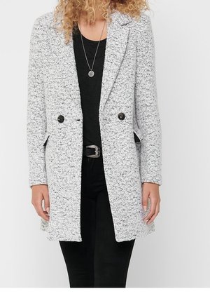 Vrouw met een gestructureerde lichtgrijze jas met dubbele knopen over een zwarte blouse en broek, aangevuld met een hangertje als ketting en een zwarte riem.