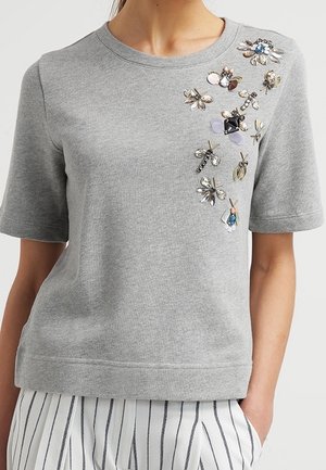 Haut gris à manches courtes orné d'insectes et de fleurs colorées en pierres précieuses sur le côté supérieur gauche, porté avec un pantalon blanc rayé.