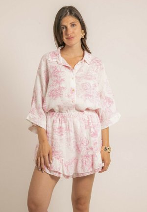 Jeune femme portant une chemise ample blanche et rose à fleurs avec un short à volants assorti, un bracelet en or et des boucles d’oreilles dorées à motif floral, debout devant un mur clair.
