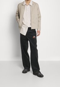 Ljusbeige bomberjacka över en krämfärgad skjorta med knappar, tillsammans med svarta byxor med en röd Nike-logotyp och svarta träningsskor.