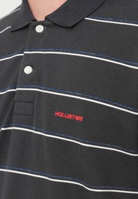 Μαύρο πόλο μπλουζάκι με λευκές και μπλε οριζόντιες ρίγες, με κλασικό γιακά και κόκκινο κεντημένο λογότυπο "Hollister" στο στήθος.