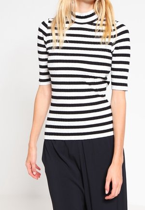 Vrouw draagt een zwart-wit horizontaal gestreept geribbeld kortärmelig shirt en losse zwarte broek, staand tegen een effen achtergrond.