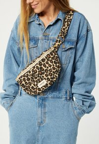 Sac besace en imprimé léopard avec une forme arrondie, de couleurs beige et noir, doté d'une fermeture éclair et d'une étiquette de marque. Bandoulière réglable visible.