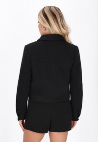 Zwarte cropped jas met een geknipt kraag, lange mouwen en knoopmanchetten, gemaakt van gladde stof. Gedragen over zwarte shorts.
