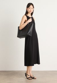 Abito nero senza maniche con una silhouette fluida che arriva a metà polpaccio. Tessuto morbido con scollatura rotonda. La modella tiene una borsa a tracolla nera con manici visibili.