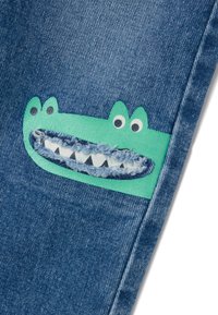 Tela de mezclilla con un tono azul que presenta un gran aplique de cocodrilo verde; incluye ojos, una boca sonriente y un detalle texturizado en los dientes.