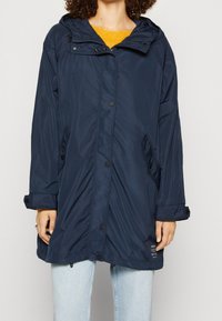 Femme portant un manteau de pluie à capuche bleu marine sur un pull jaune et un jean bleu clair, avec fermeture à boutons et à zip à l'avant.