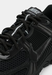 Μαύρο αθλητικό παπούτσι Nike Vomero 5 με άνω μέρος από δίχτυ, γυαλιστερές λεπτομέρειες, υφασμένα πλαϊνά πάνελ, επίπεδα κορδόνια και ανακλαστικές λεπτομέρειες.
