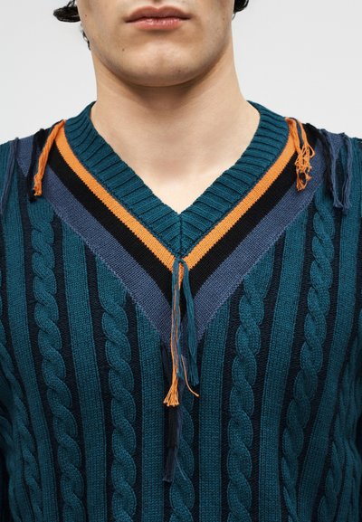Homme portant un pull en maille torsadée bleu sarcelle avec un col en V rayé orange, noir et bleu, orné de franges lâches en fil au niveau des bords du col.