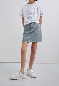 Enfant portant un t-shirt blanc avec un design de skull brodé, une jupe en imprimé zèbre vert et des baskets blanches à velcro, se tenant sur un sol gris clair.